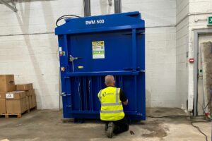 A man in hi-viz bent down beside a blue waste baler